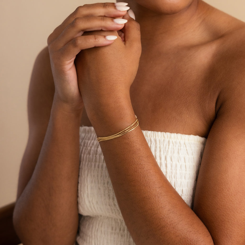 A woman in a white textured top wears the Triple Snake Chain Bracelet, her minimal jewelry choice highlighted as she gently clasps her hands near her face, creating a refined and elegant layered style.