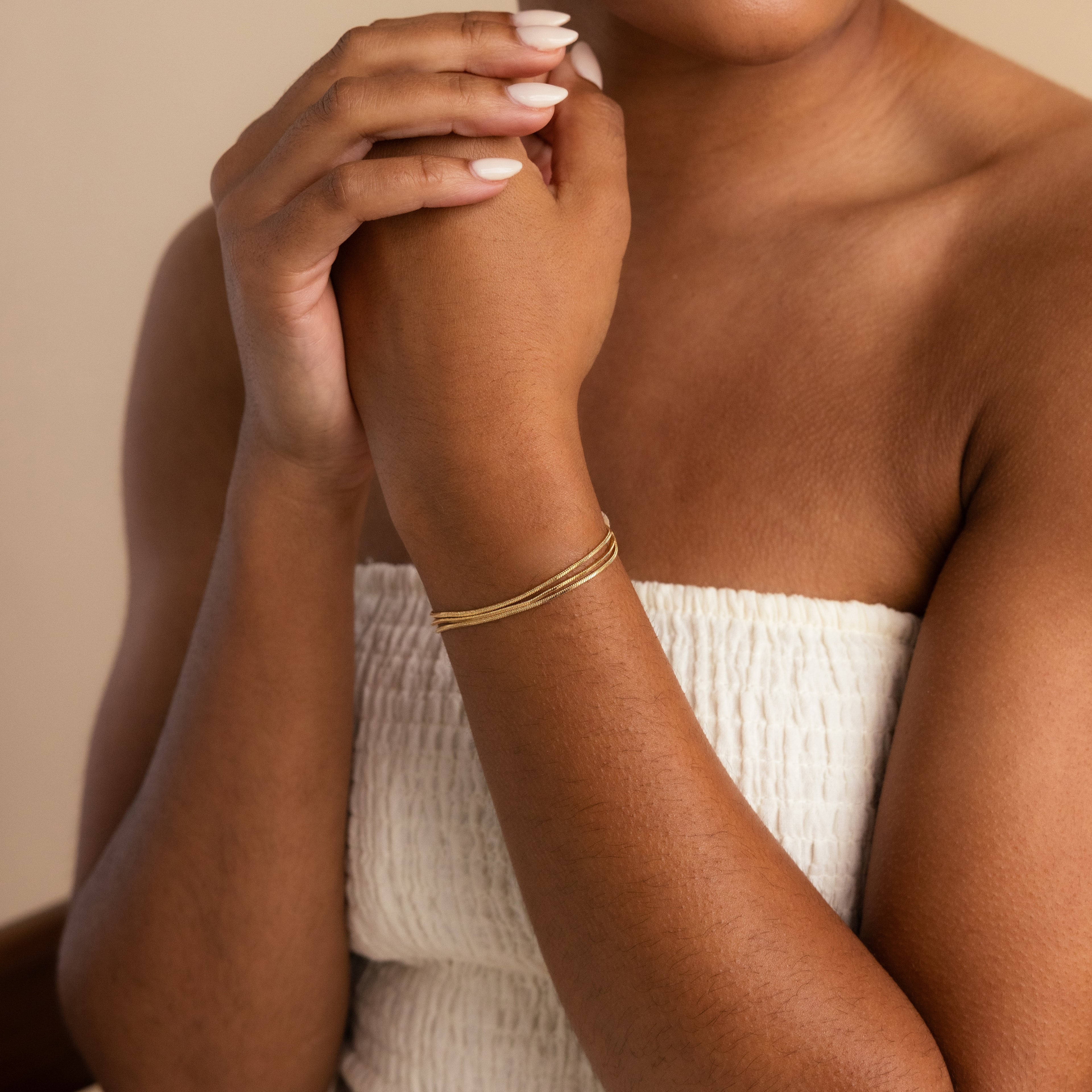 A woman in a white textured top wears the Triple Snake Chain Bracelet, her minimal jewelry choice highlighted as she gently clasps her hands near her face, creating a refined and elegant layered style.