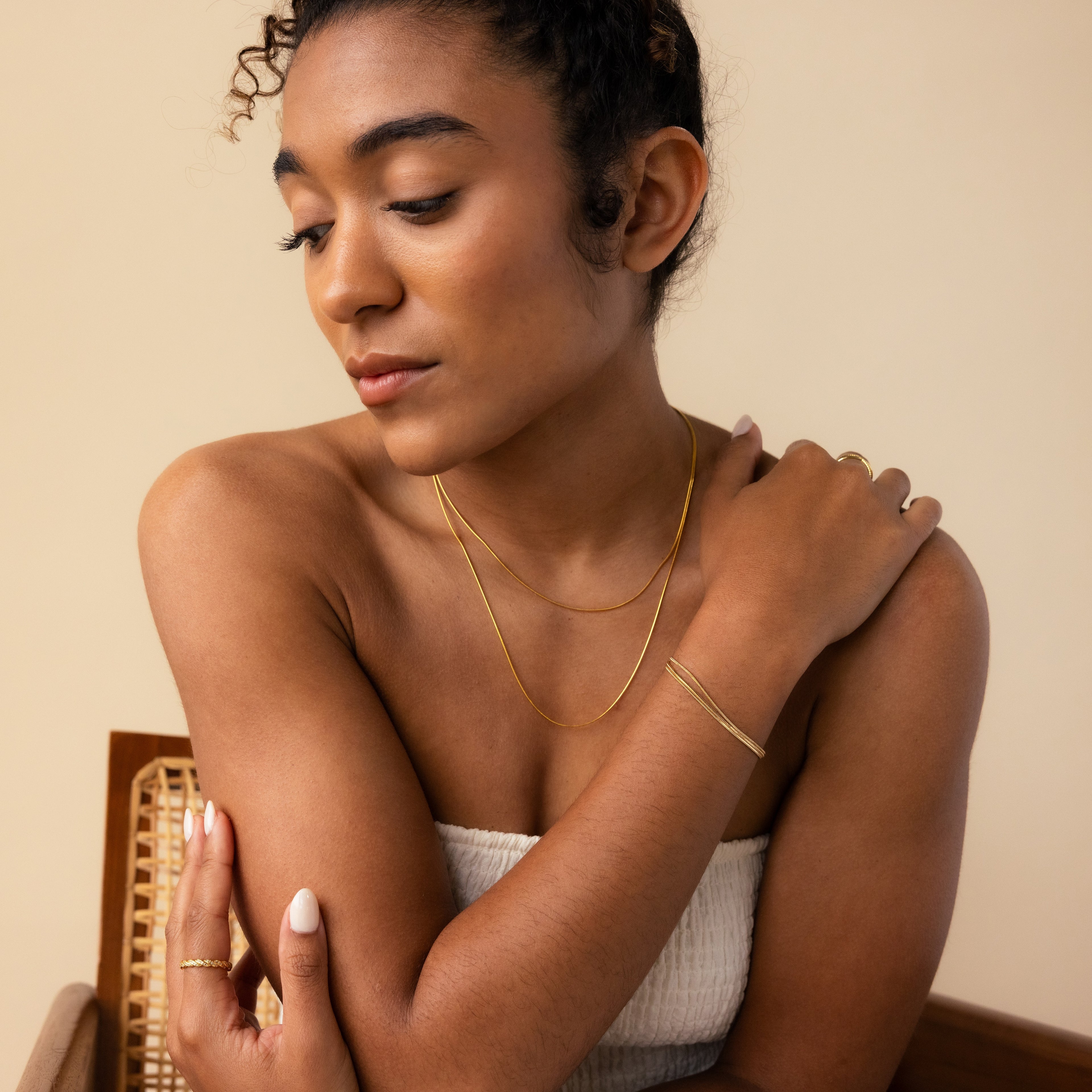 A woman in a white top poses gracefully, wearing the Triple Snake Chain Bracelet in White Gold as minimal jewelry and gazing downward with a serene expression.