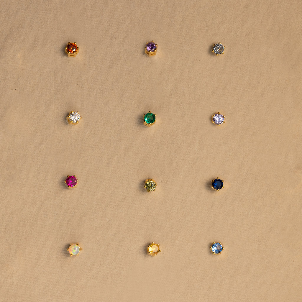 Twelve dainty earrings, including Garnet Flatback Studs in various colors, are arranged in a grid on a beige background.