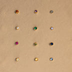 Twelve dainty earrings, including Garnet Flatback Studs in various colors, are arranged in a grid on a beige background.