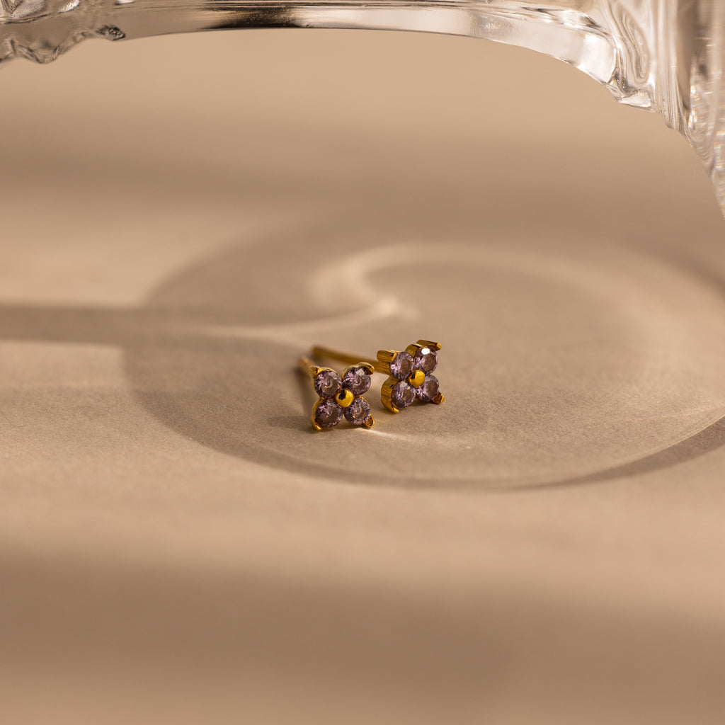A pair of gold-toned stud earrings with four small light purple gemstones arranged in a square, resting on a beige surface with a soft shadow and a glass object partially visible at the top.