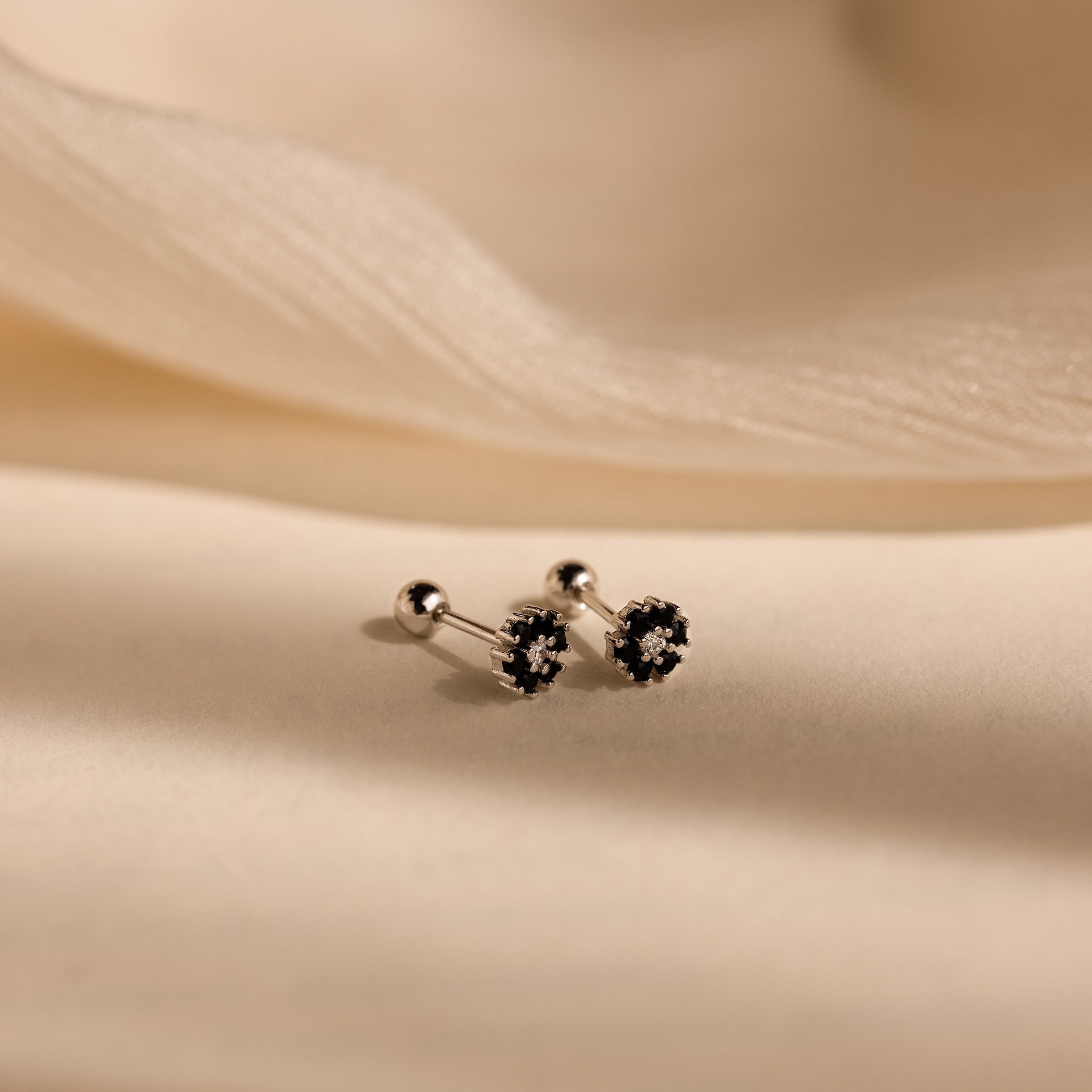 The Onyx Flower Studs are dainty silver earrings featuring dark onyx stones in a floral design, displayed against a soft beige fabric background.