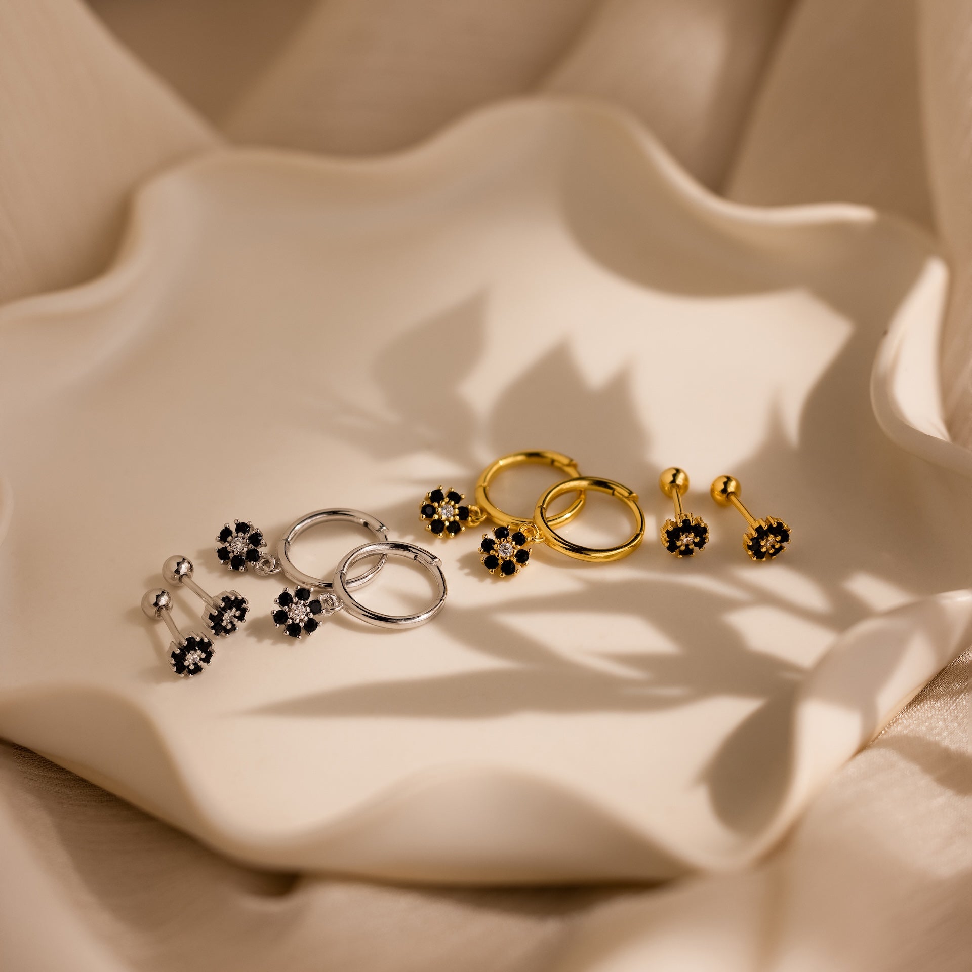 Three pairs of floral earrings, including Onyx Flower Studs in Silver and gold diamond center earrings, are displayed on a wavy-edged cream ceramic plate.