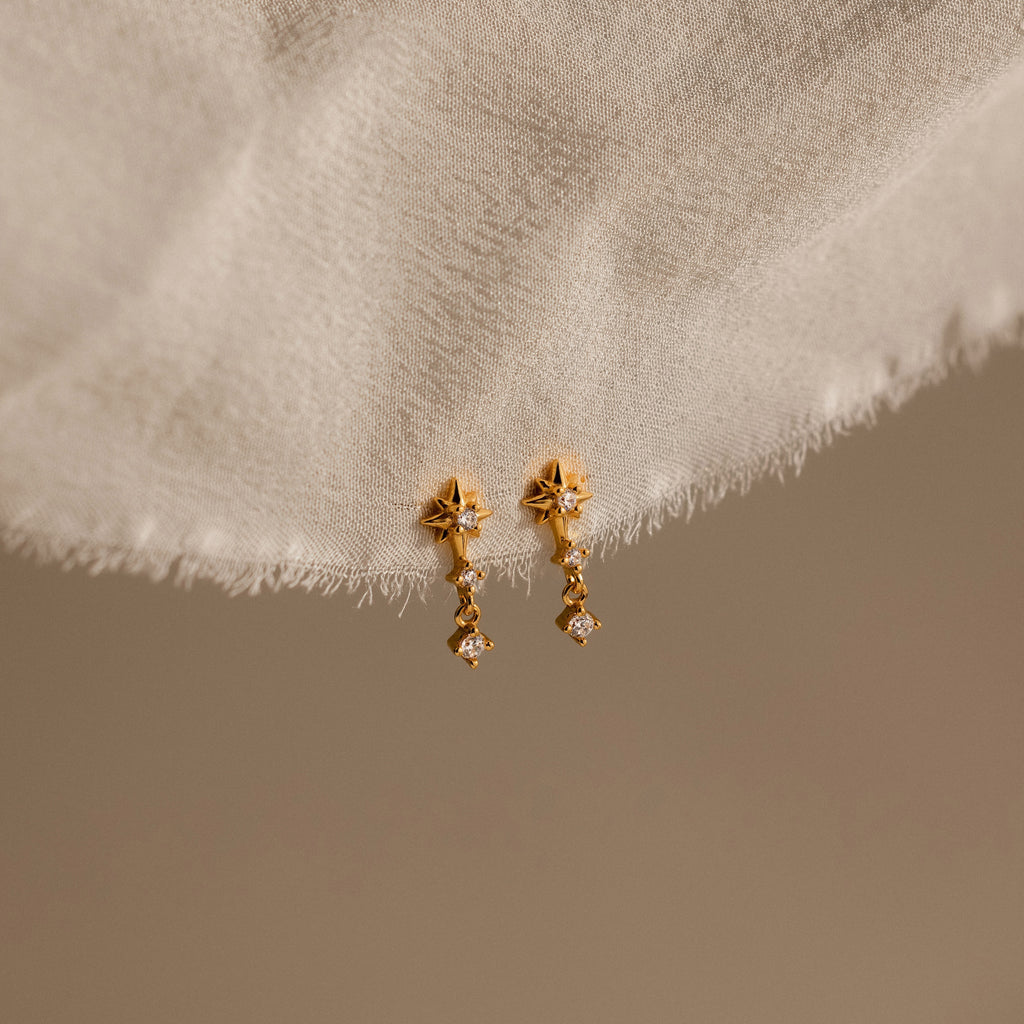 Diamond Starburst Drop Studs in 18K Gold, featuring gold star-shaped drops with small dangling crystals, elegantly displayed on soft cream-colored fabric.