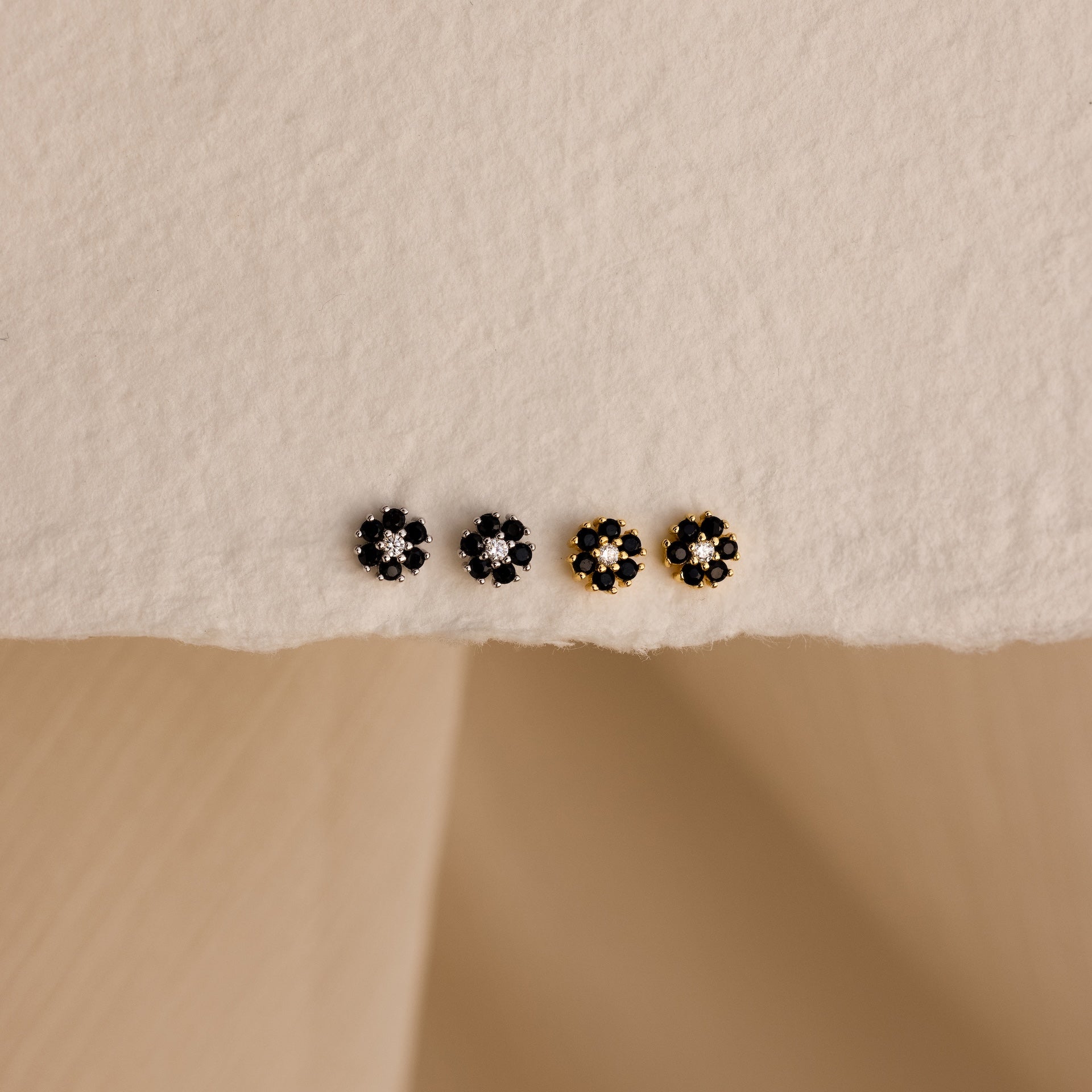 A set of four Onyx Flower Studs—two black onyx and two gold flower-shaped earrings—arranged on a textured cream surface.