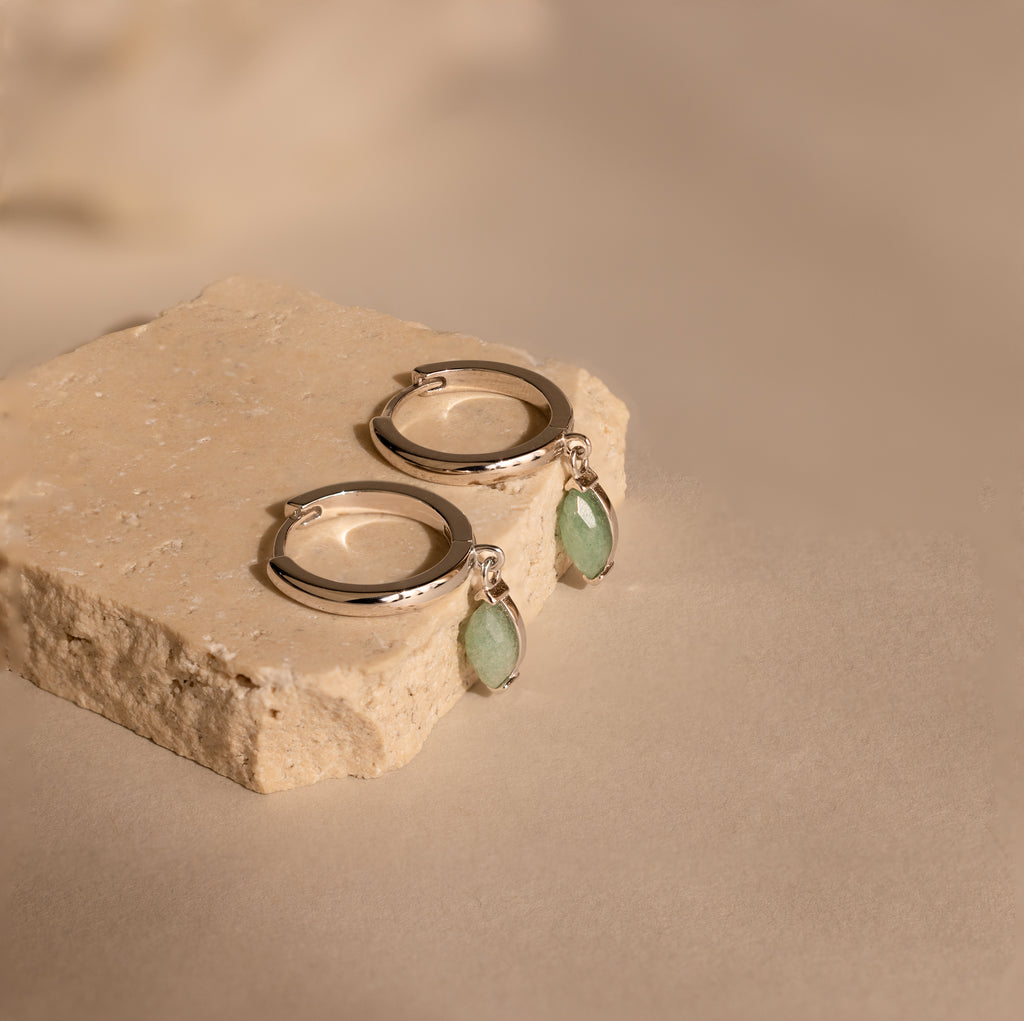 The Jade Marquise Huggies in Silver, featuring green stone charms, are displayed on a beige stone block against a neutral background, highlighting their elegant design.