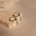 The Jade Marquise Huggies in Silver, featuring green stone charms, are displayed on a beige stone block against a neutral background, highlighting their elegant design.