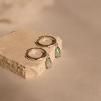 The Jade Marquise Huggies in Silver, featuring green stone charms, are displayed on a beige stone block against a neutral background, highlighting their elegant design.
