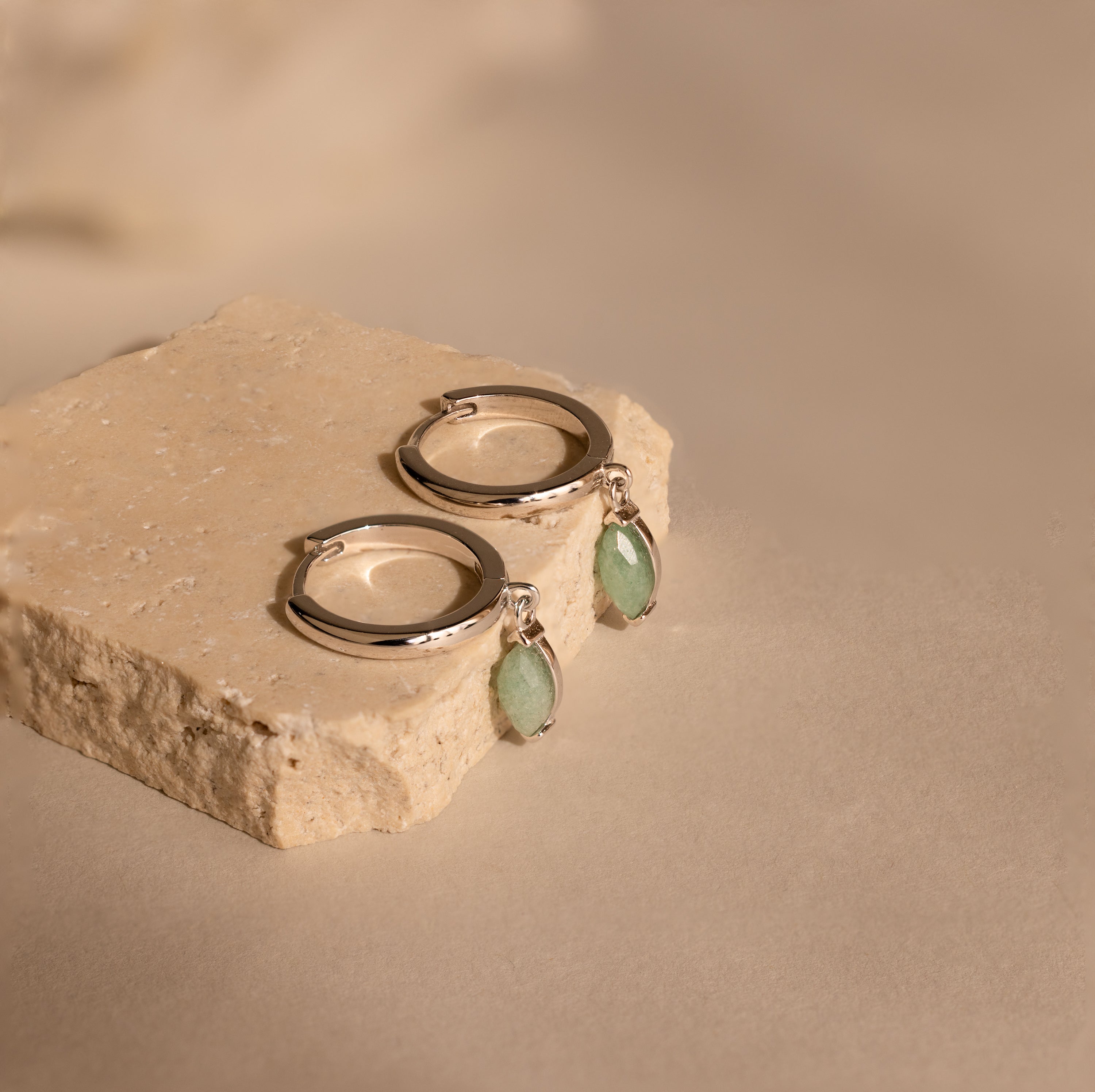 The Jade Marquise Huggies in Silver, featuring green stone charms, are displayed on a beige stone block against a neutral background, highlighting their elegant design.