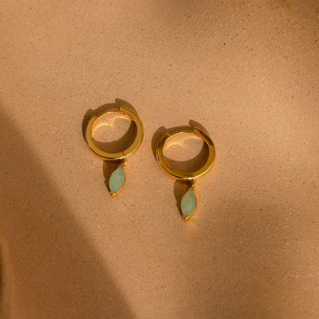 Jade Marquise Huggies in 18K Gold with green marquise stones are displayed on a sandy beige background.