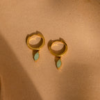 Jade Marquise Huggies in 18K Gold with green marquise stones are displayed on a sandy beige background.