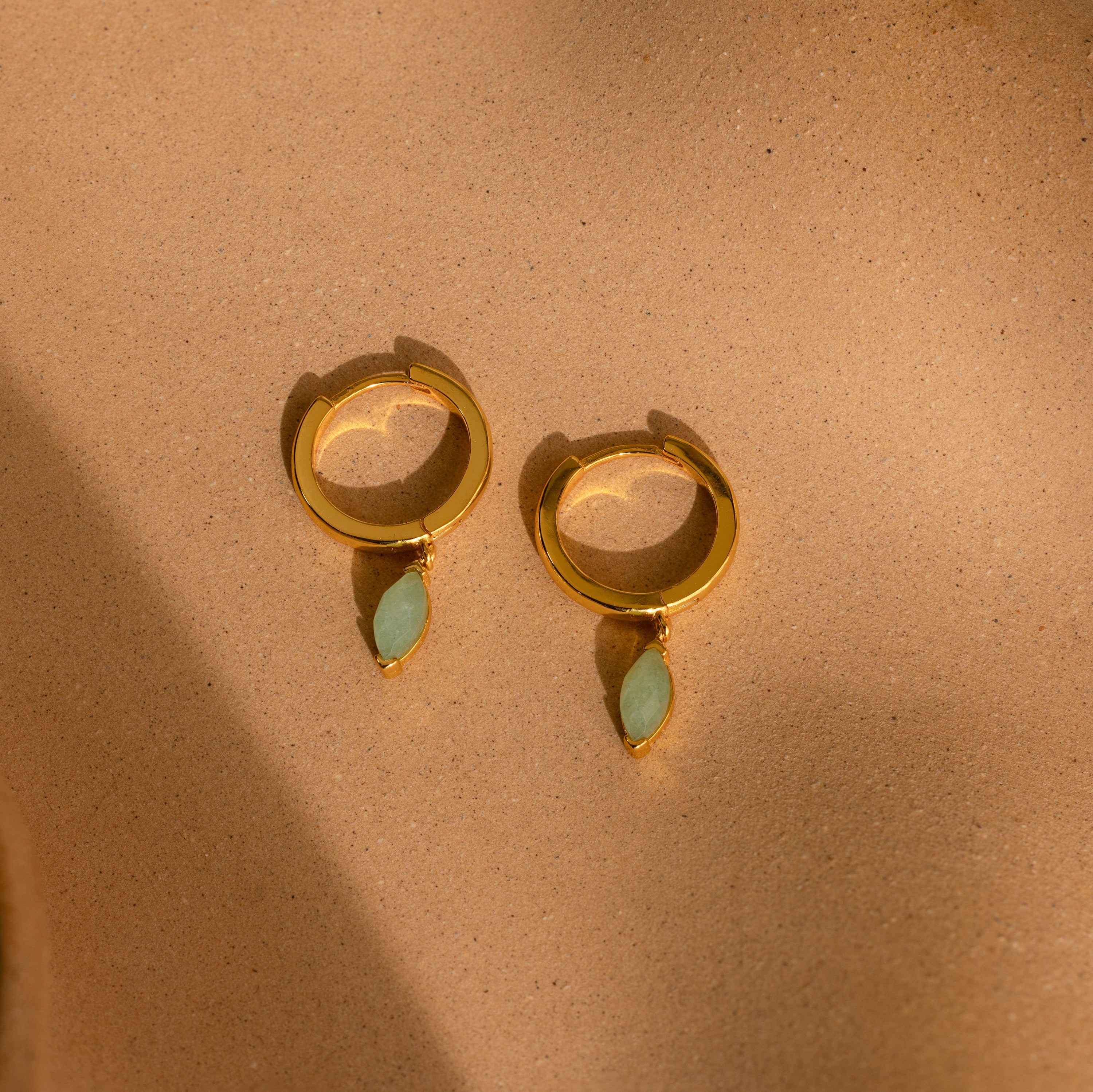 Jade Marquise Huggies in 18K Gold with green marquise stones are displayed on a sandy beige background.