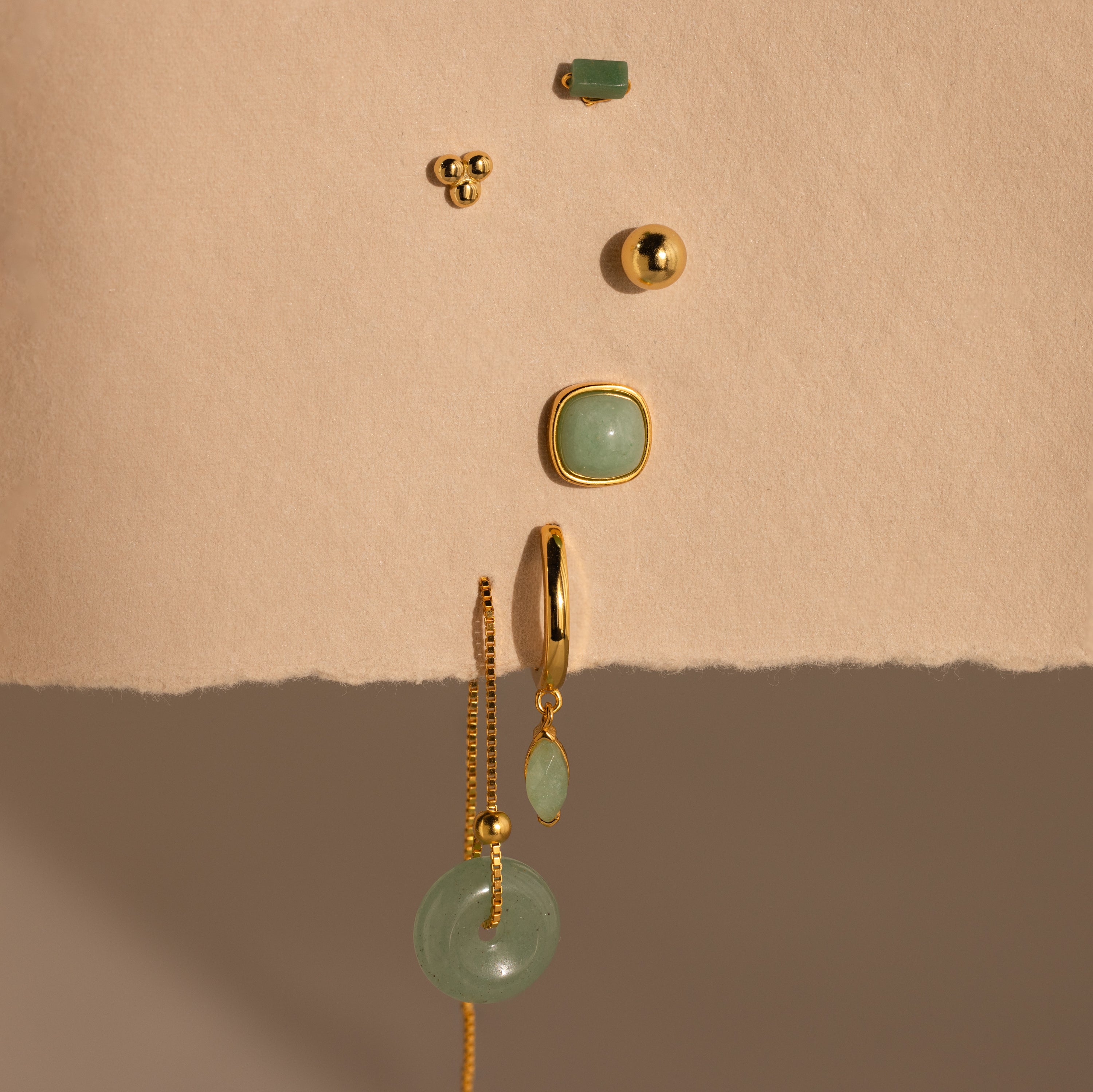 Jade Marquise Huggies, elegant gold and green earrings, displayed vertically on a textured beige background.