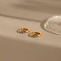 Rhombus Blue Zircon Huggies—these gold hoop earrings with square blue zircon stones are shown on a beige surface with soft shadows and glass nearby.