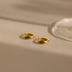 The Rhombus Peridot Huggies feature gold hoops with green rectangular stones on a beige surface, casting a soft shadow. These gemstone huggie earrings have glass details and are perfect, customizable accessories for any occasion.