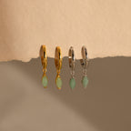 Two pairs of Jade Marquise Huggies in gold and silver, featuring green marquise jade stones, are displayed on a beige textured surface.