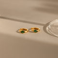 Rhombus Emerald Huggies—customizable gold hoop earrings with green rhombus gemstones—rest on a beige surface, softly illuminated by natural light.