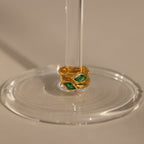 Two Rhombus Emerald Huggies with green stones are stacked around the stem of a clear wine glass placed on a beige surface.