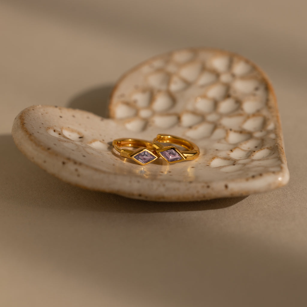 A pair of gold Rhombus Alexandrite Huggies with purple gemstones sits on a textured, heart-shaped ceramic dish—beautiful customizable earrings for any occasion.