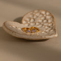 A pair of gold Rhombus Alexandrite Huggies with purple gemstones sits on a textured, heart-shaped ceramic dish—beautiful customizable earrings for any occasion.
