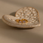 A pair of gold Rhombus Alexandrite Huggies with purple gemstones sits on a textured, heart-shaped ceramic dish—beautiful customizable earrings for any occasion.
