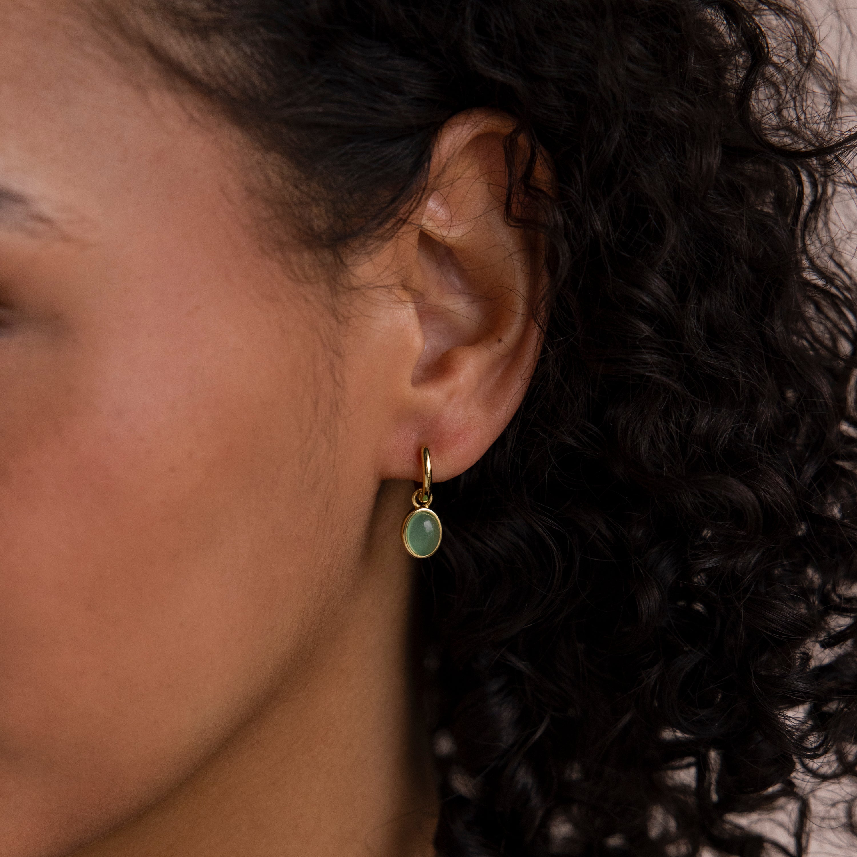 Close-up of a person’s ear adorned with Jade Drop Hoops—gold huggie earrings featuring a small oval jade gemstone, with curly hair visible.