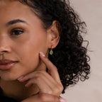A woman with curly hair touches her face, wearing Jade Drop Hoops in 18K Gold adorned with a green jade gemstone, set against a neutral background.