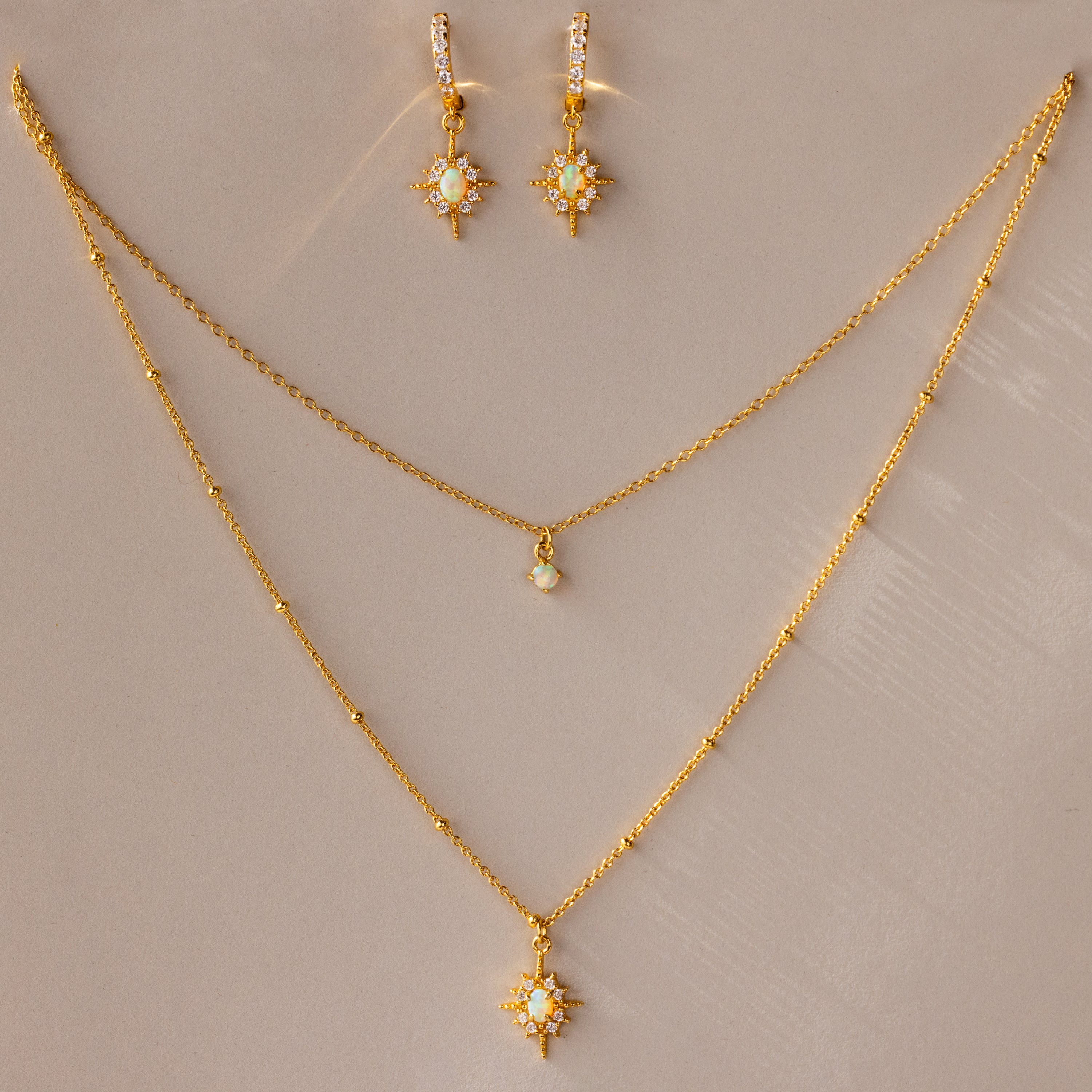 The Opal Star Necklace & Earrings Set includes a gold-layered necklace with opal star pendants and matching star-shaped earrings, beautifully displayed on a beige surface.