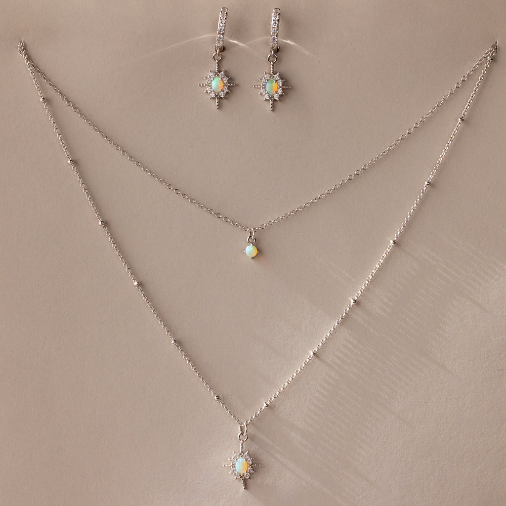 The Opal Star Necklace & Earrings Set in Silver features shimmering opal and crystal starburst pendants, beautifully displayed on a beige background.