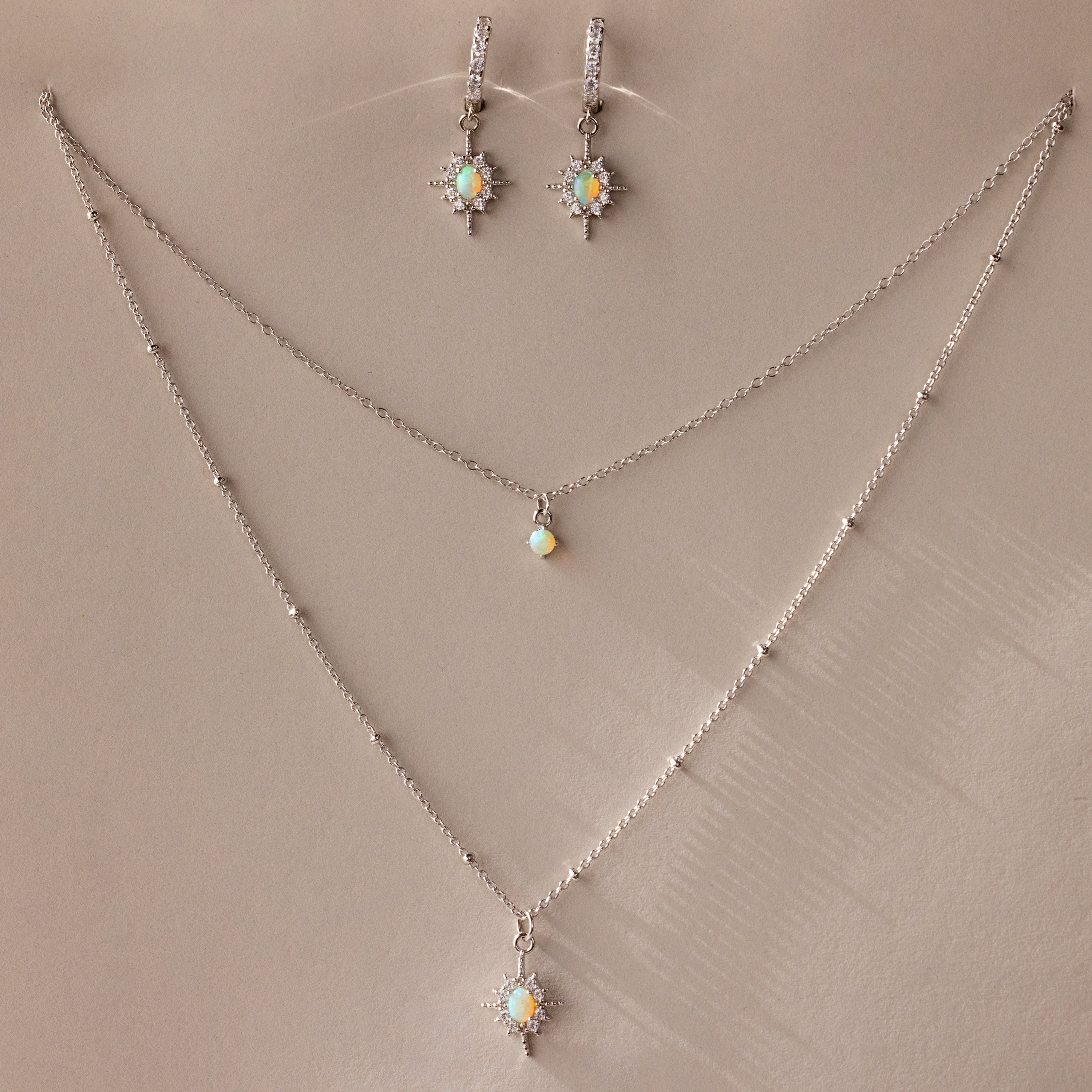 The Opal Star Necklace & Earrings Set in Silver features shimmering opal and crystal starburst pendants, beautifully displayed on a beige background.