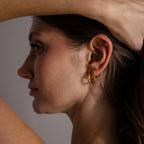 A woman with brown hair and gold earrings lifts her arm, showing off her ear adorned with the Beaded Hoops in 18K Gold jewelry.