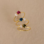 Three Birthstone & Pearl Rings with red, blue, and green gemstones plus pearls are arranged on beige. Each Birthstone & Pearl Ring is ideal for those who love elegant, personalized jewelry.