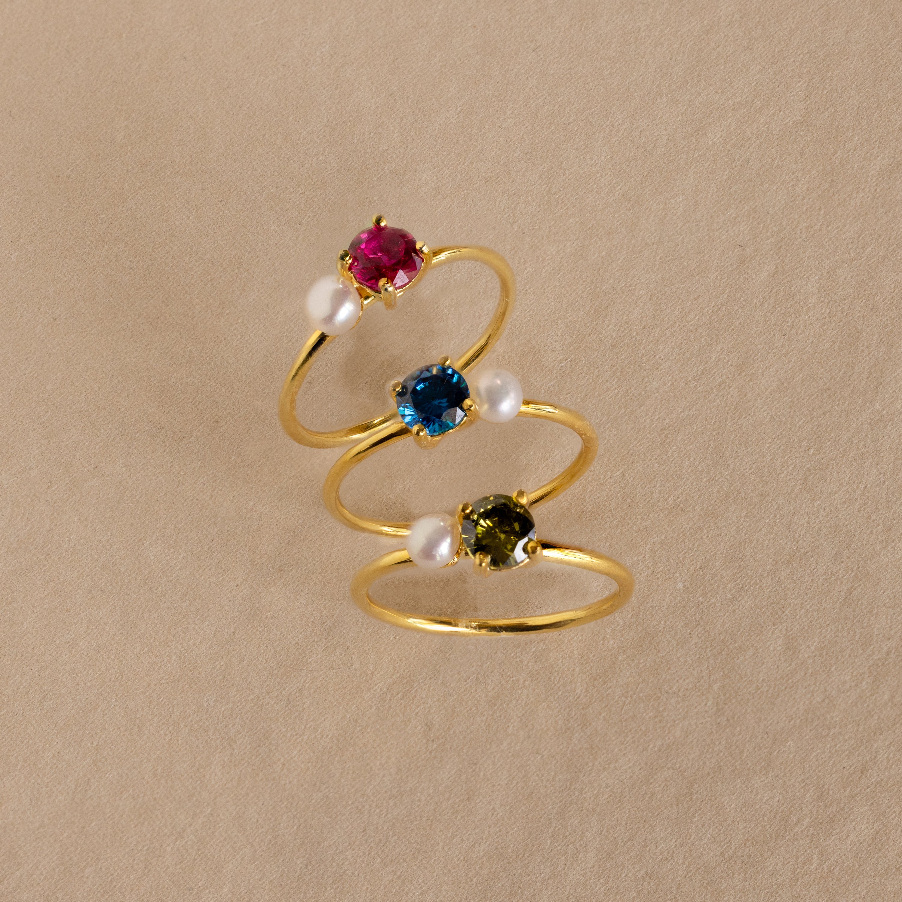 Three Birthstone & Pearl Rings with red, blue, and green gemstones plus pearls are arranged on beige. Each Birthstone & Pearl Ring is ideal for those who love elegant, personalized jewelry.