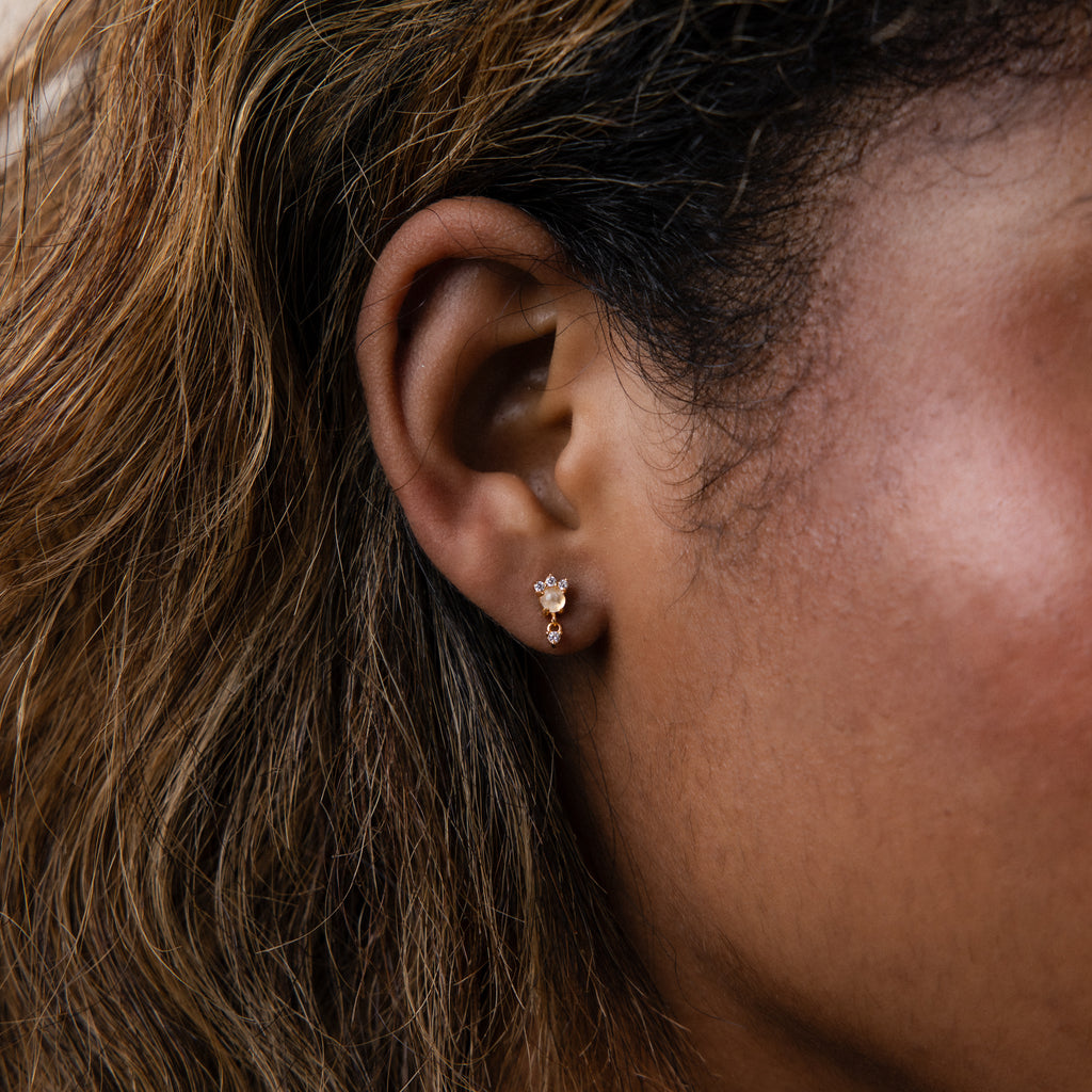 Close-up of a woman's ear wearing the Halo Moonstone Drop Studs in 18K Gold, featuring three delicate stones for a touch of royal elegance.