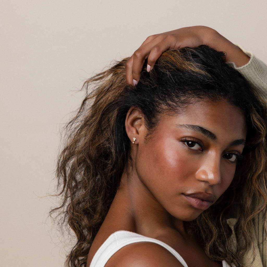 Woman with curly hair touches her head, wearing Halo Moonstone Drop Studs and a sleeveless top, looking at the camera against a neutral background.