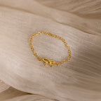 A delicate gold chain ring with a small bow centerpiece displayed on a soft, sheer beige fabric.