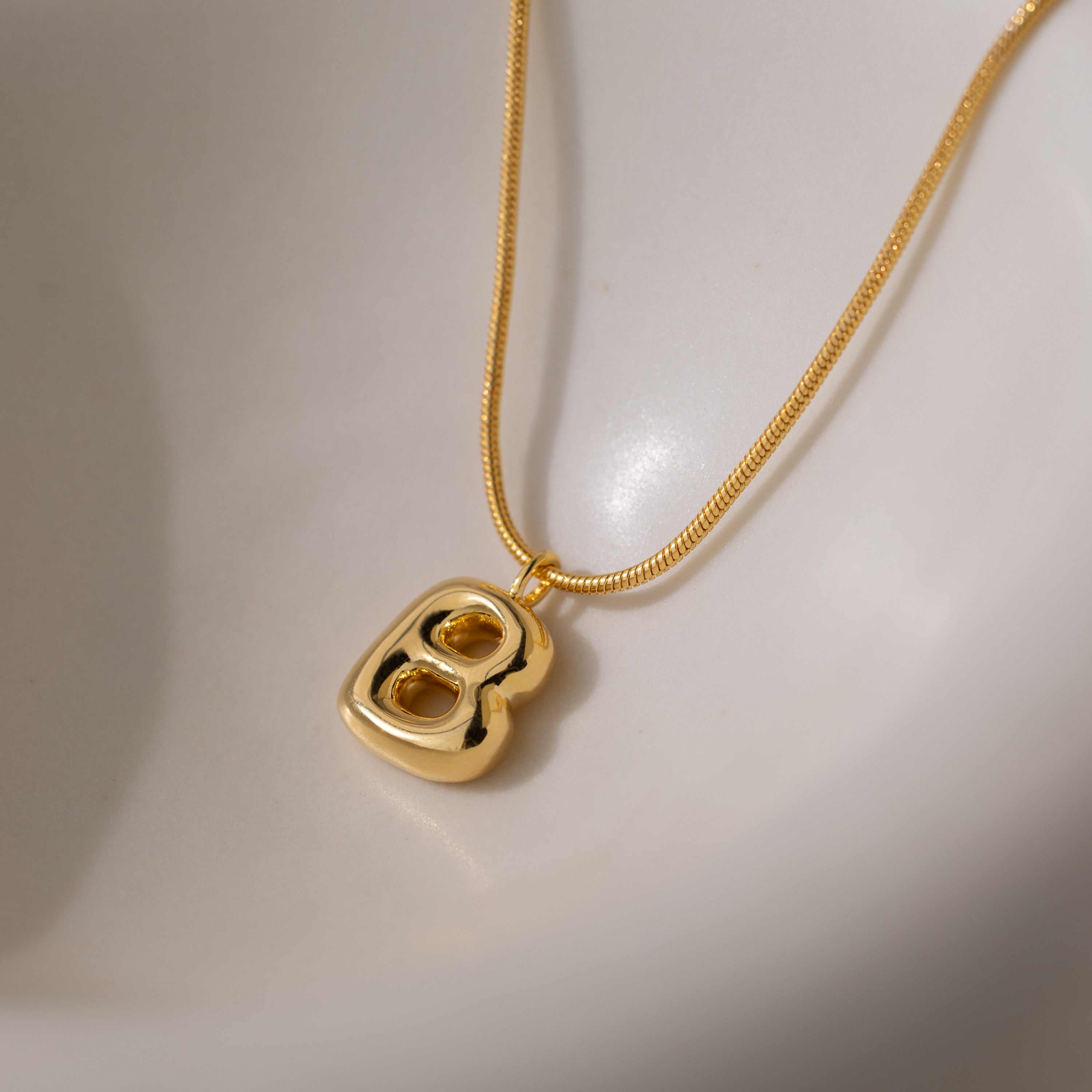 The Bubble Letter Necklace in 18K Gold features a bold, shiny letter B charm on a smooth white surface—ideal for anyone who loves personalized jewelry.