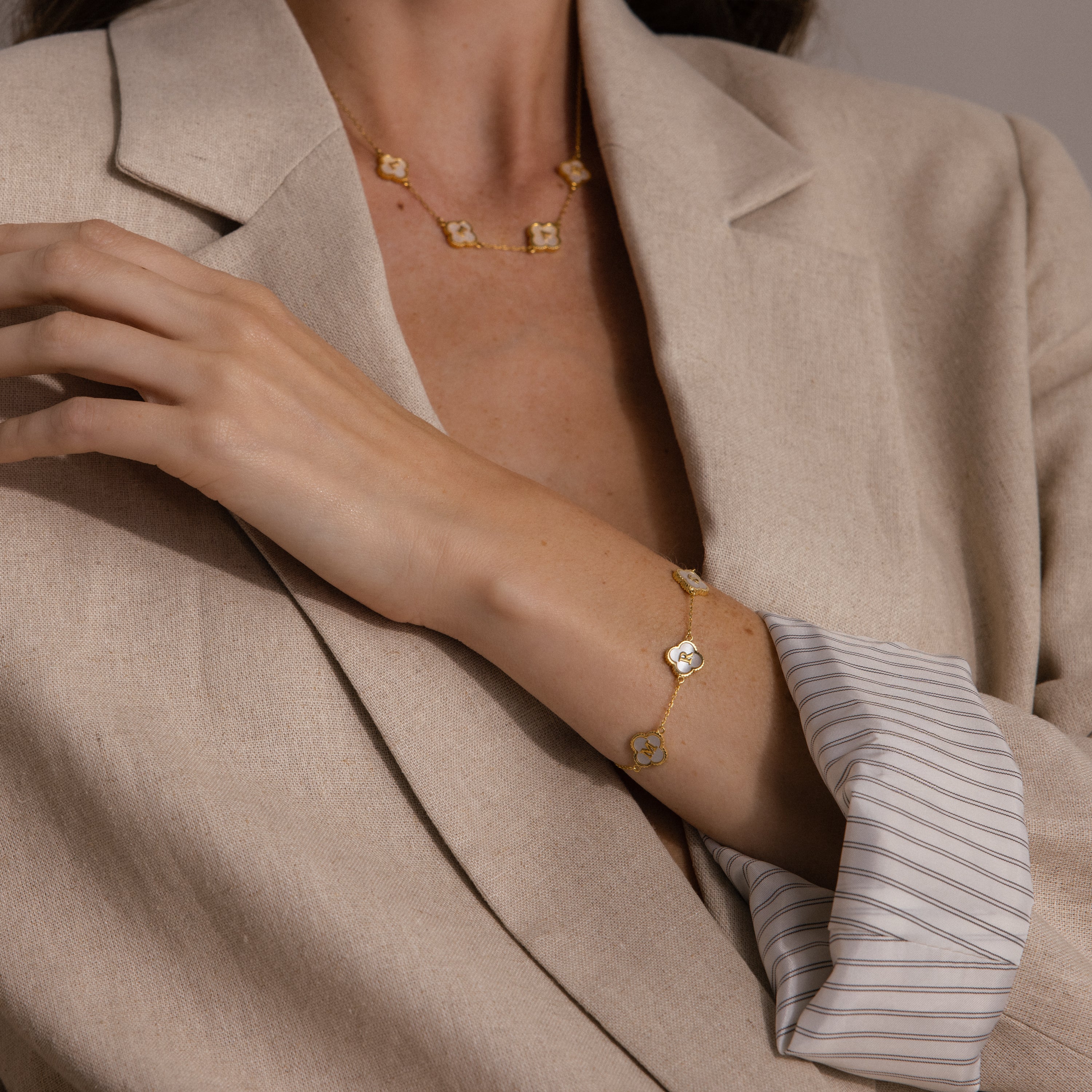 Woman in a beige blazer wearing a gold necklace and the Pearl Clover Letter Bracelet adorned with matching mother of pearl clovers.