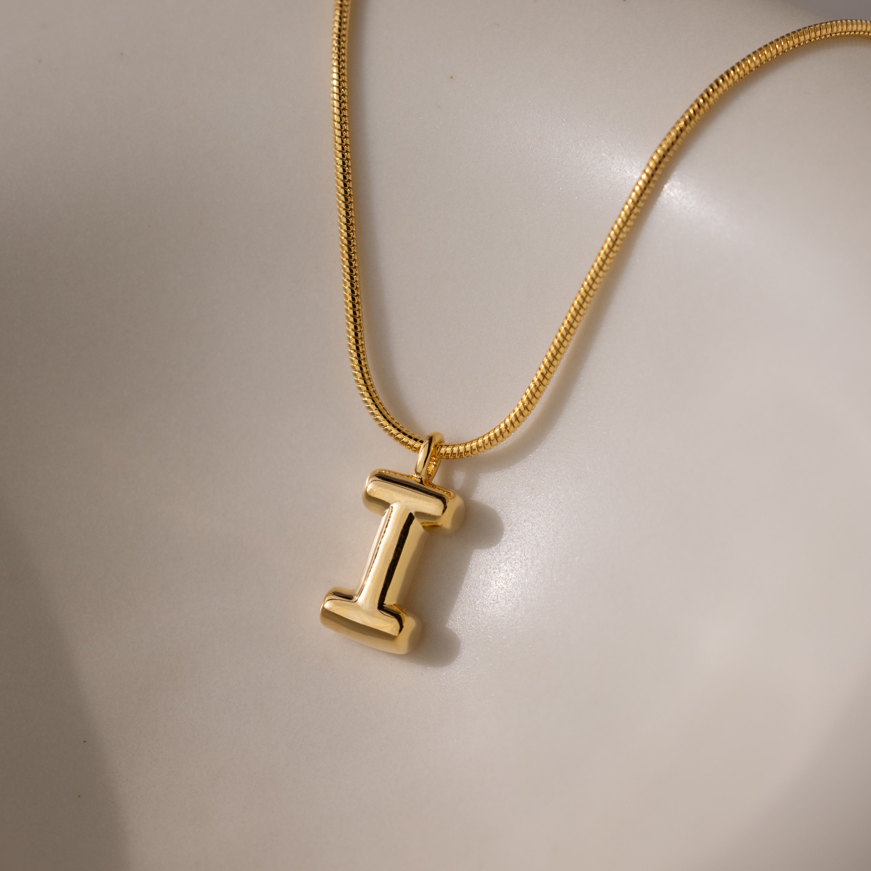 Bubble Letter Necklace in 18K Gold featuring a chunky letter I charm, displayed on a smooth, light-colored surface.