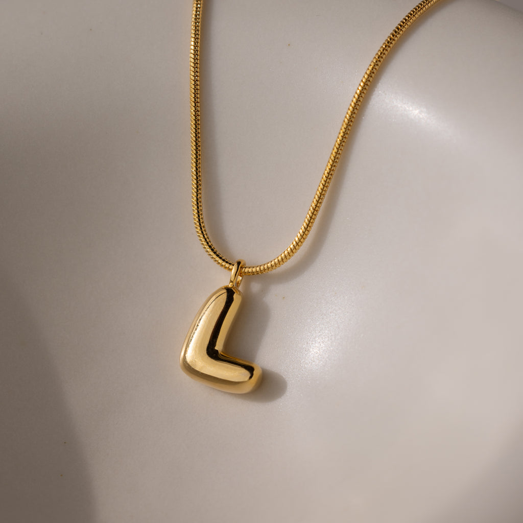 The Bubble Letter Necklace in 18K Gold, featuring a chunky L-shaped charm, rests on a smooth off-white surface.