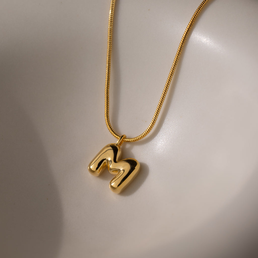 The Bubble Letter Necklace in 18K Gold features a shiny M pendant resting on a smooth, light-colored surface—an ideal personalized jewelry piece.
