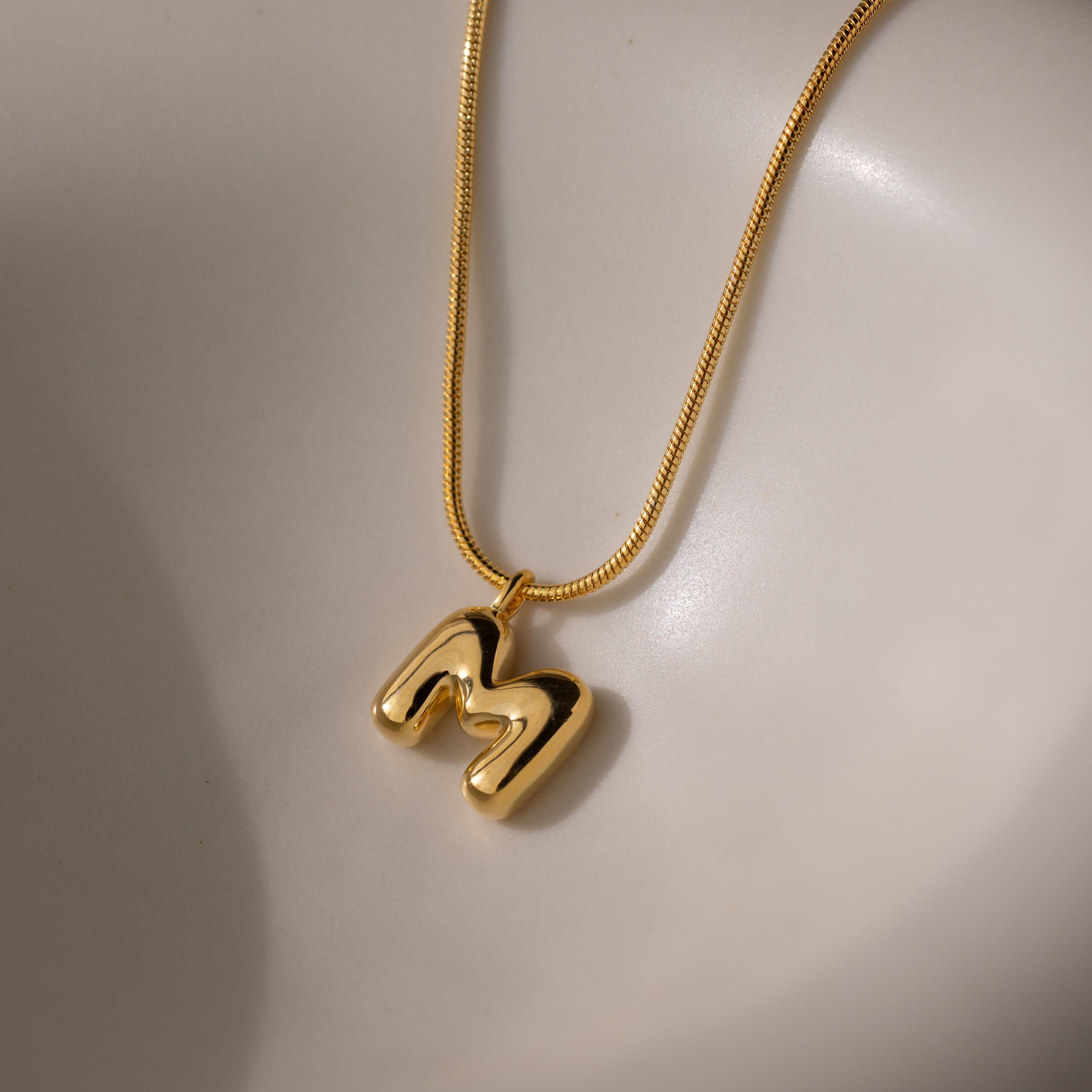 The Bubble Letter Necklace in 18K Gold features a shiny M pendant resting on a smooth, light-colored surface—an ideal personalized jewelry piece.