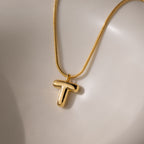 The Bubble Letter Necklace in 18K Gold features a shiny, bold letter T pendant in a chunky charm style, displayed on a smooth, light surface.