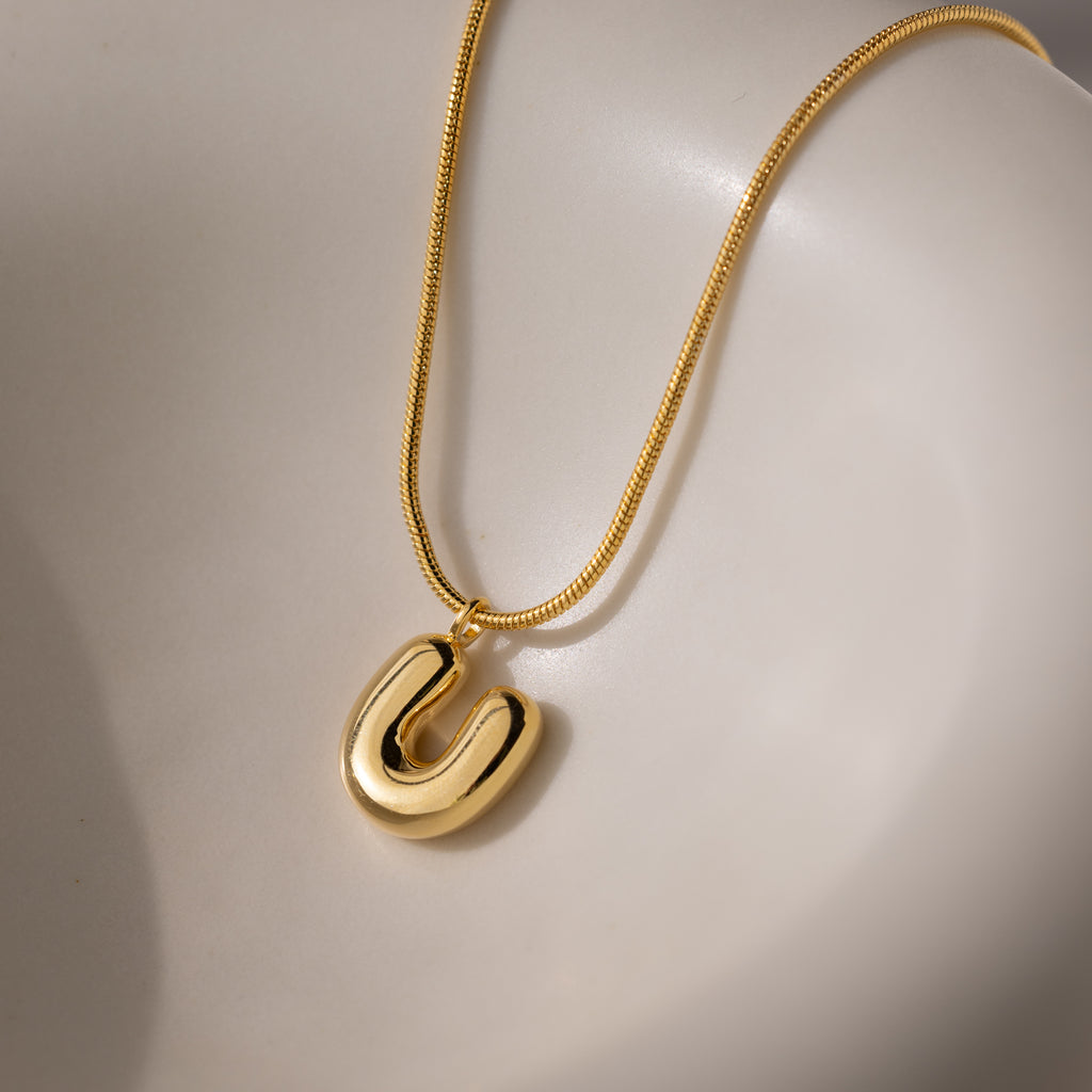 The Bubble Letter Necklace in 18K Gold features a U-shaped pendant and is displayed on a smooth, light-colored surface.