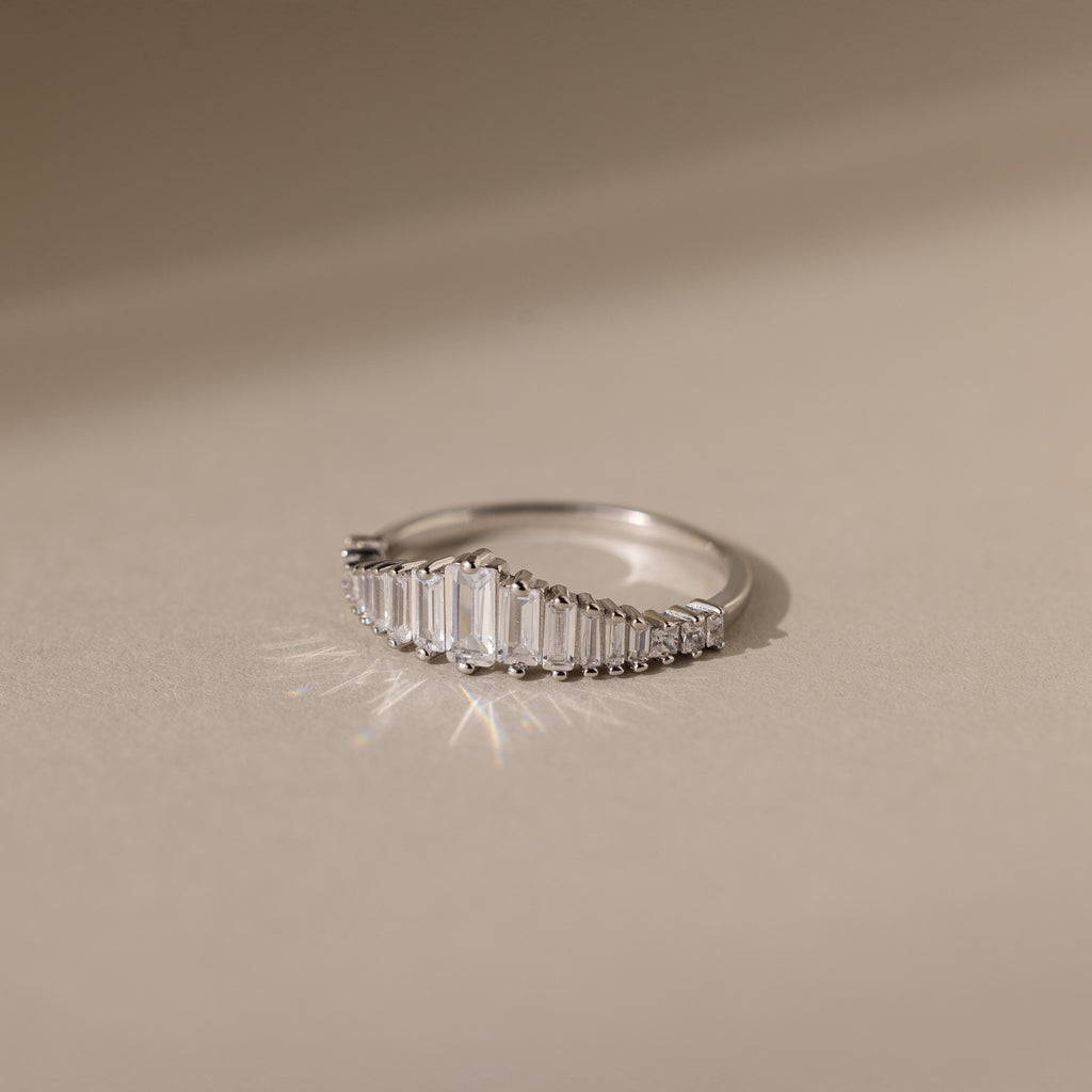 The Art Deco Diamond Ring in Sterling Silver features rectangular diamonds set in a curved line, casting a soft shadow on a beige surface—a striking statement piece.