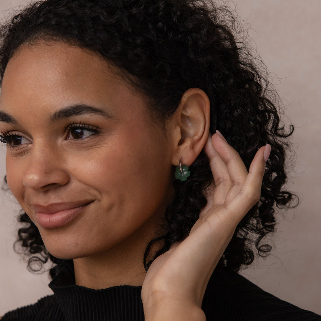 A woman with curly hair smiles as she touches her ear, showing off Jade Flower Hoops—a meaningful and dainty gift.