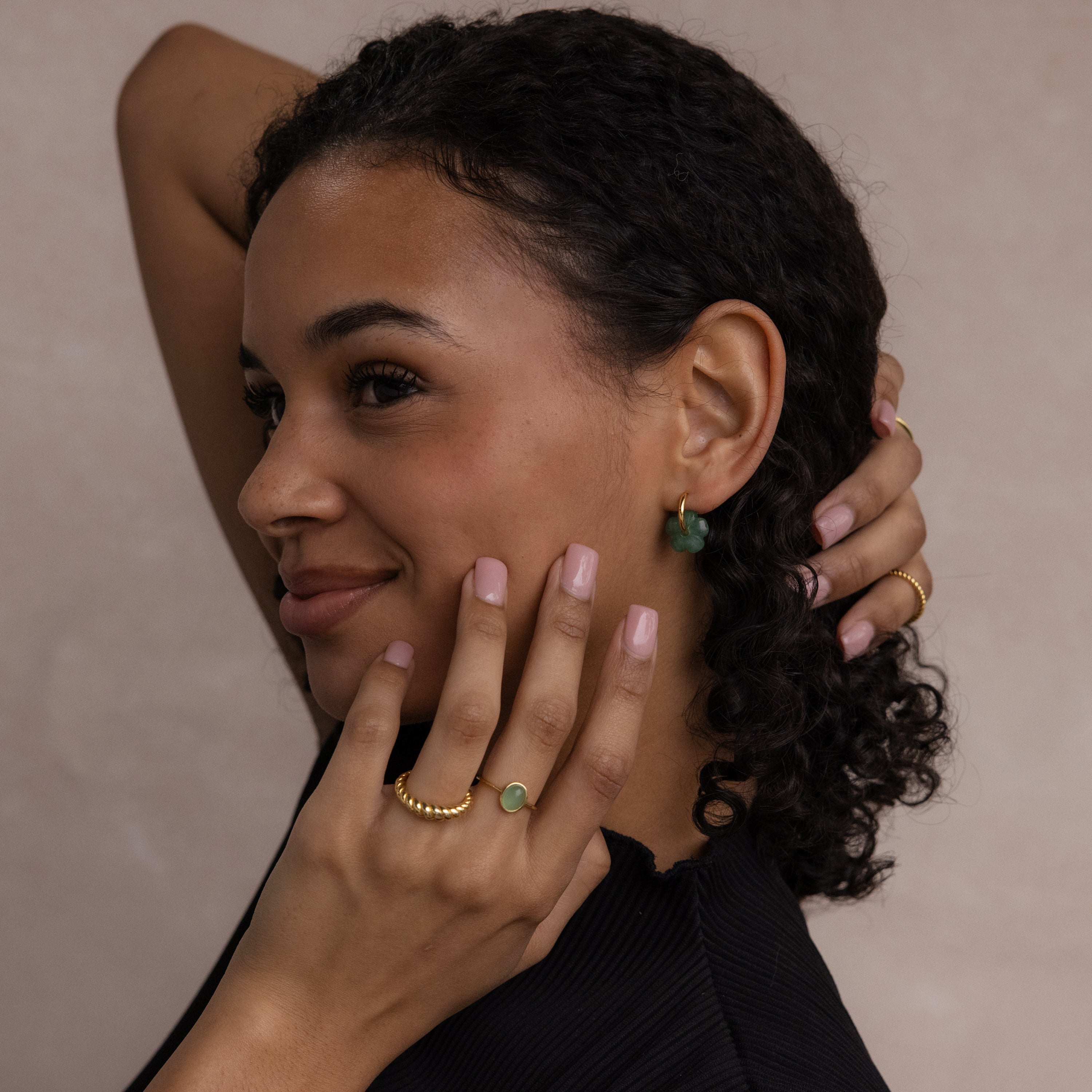 A woman with curly hair wears Jade Flower Hoops—delicate gold and green earrings—holding her hand to her face and smiling, showcasing these meaningful earrings as the perfect gift.