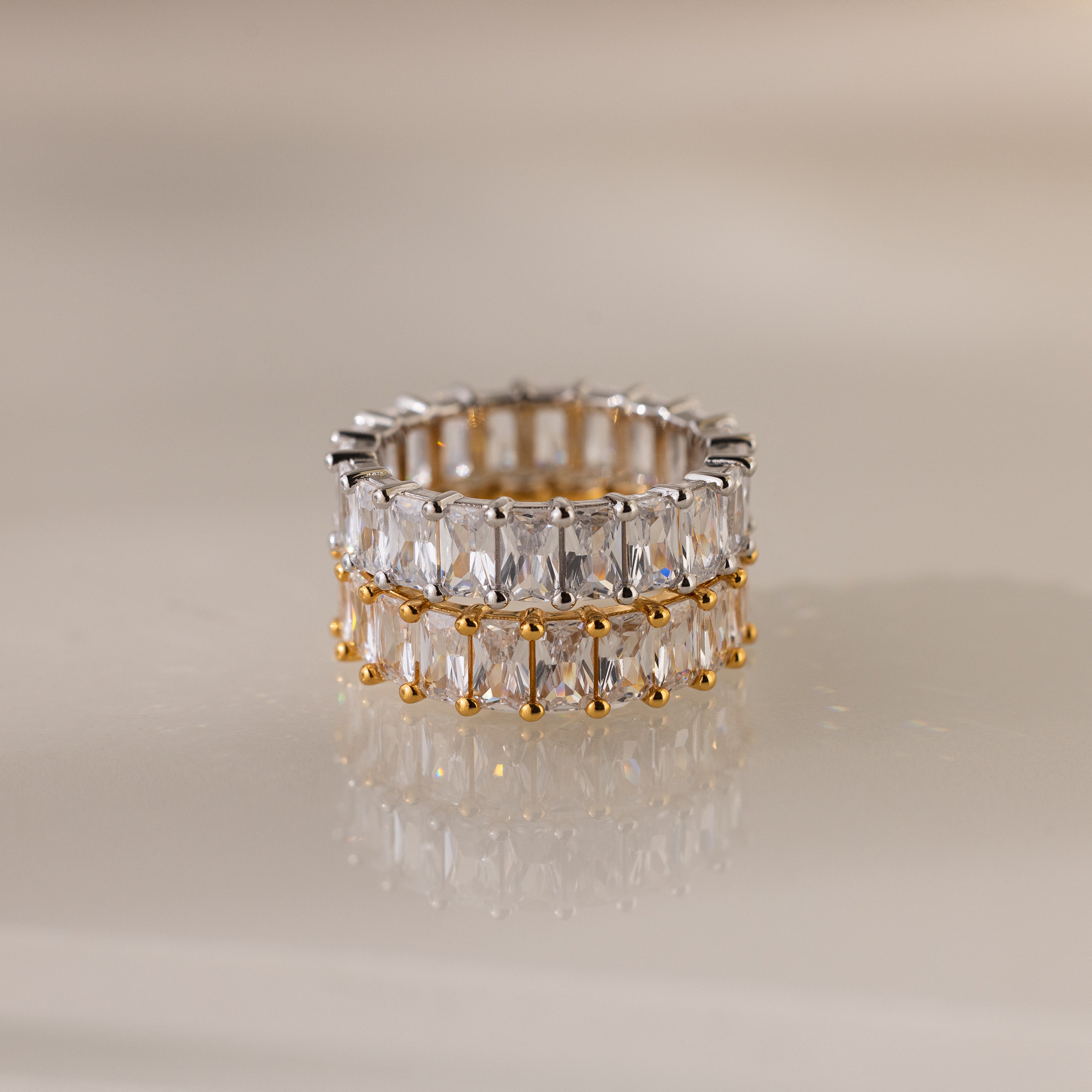 The Colette Eternity Ring features gold and silver bands with dual rows of baguette diamonds, exuding opulence against a neutral backdrop.
