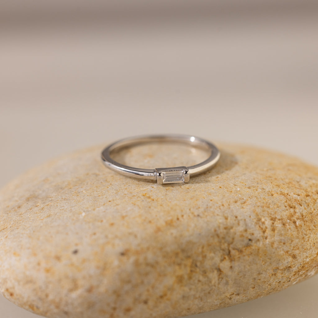 The Baguette Diamond Ring in Silver features a dainty baguette diamond in an east-west bezel setting, displayed on a smooth, light-colored stone.